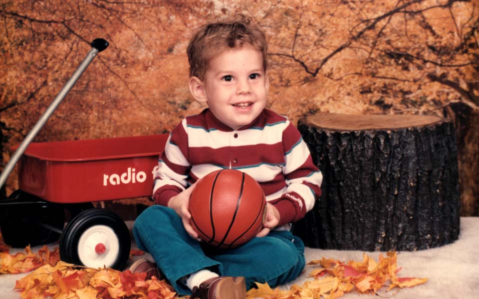 Jason as an adorable toddler holding a basketball in front of a red radio flyer wagon amidst a obviously fake wooded backdrop.