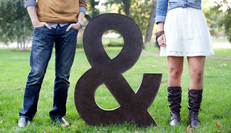 A young couple between a large metal ampersand sign artistically cropped to only show their wardrobe.