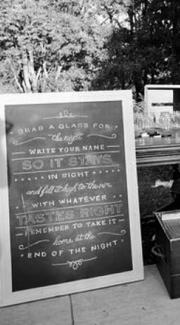 An outdoor bar with large rustic chalkboard in the foreground instructing guests to take a mason jar for the cocktail hour.